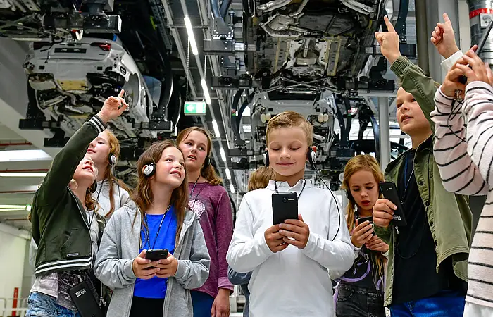 Eine Kindergruppe ausgestattet mit einem Audioguide erleben eine Werkbesichtigung bei AUDI. Die Gruppe steht unter einem Auto und betrachtet das Fahrzeug sehr interessiert.