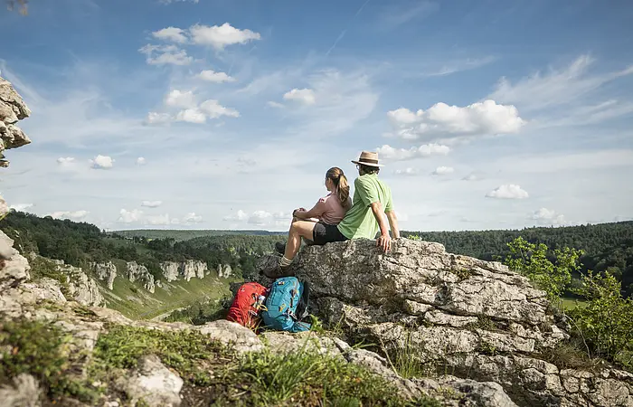 Zwei Wanderer sitzen auf einem Felsen und blicken auf bewaldete Hügel unter blauem Himmel mit Wolken.