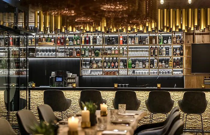 Die Bar nimmt den größten Bereich ein mit dem offenen, gut gefülltem Regal. Davor stehen dunkle bequeme Bistrostühle. Ein Tisch mit Kerzen ist noch zum Teil zu sehen.