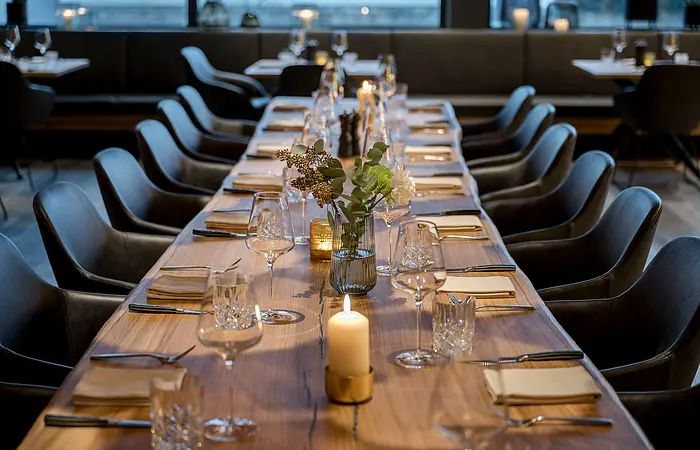 Eine lange Tafel mit Weingläser, Besteck und brennenden Kerzen ist im Mittelpunkt. Die gemütlichen schwarzen Stühlen. Im Hintergrund ist eine große Fensterfront.