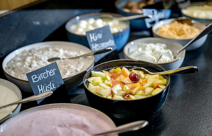 Schwarze Schüsseln gefüllt mit Bircher Müsli, Erdbeerjoghurt, Obstsalat und Quark angerichtet auf einem Tisch.