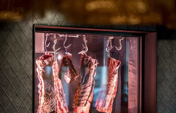 Im Restaurant ist ein Fenster in der Wand eingebaut. Durch dieses kann man einen Blick in den Kühlraum der Metzgerei mit Rinderhälften werfen. Diese reifen hier weiter. Ein Tisch steht genau vor dem Fenster und eine Sitzbank mit grauem Stoff ist an der Wand befestigt.