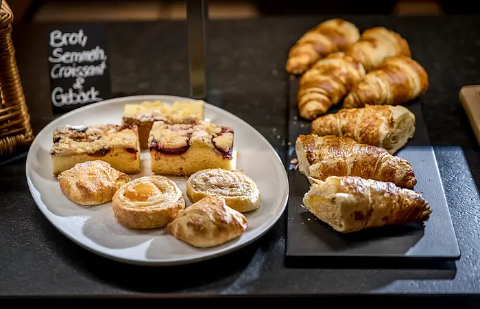 Auf schwarzen Granitplatten sind Croissants angerichtet und auf einem weißen Teller daneben befinden sich lauter kleine süße Teilchen. Dahinter ist die übliche Tafel mit der Beschriftung zu sehen.