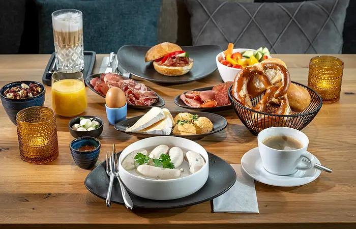 Der braune Tisch ist mit Kerzen, Orangensaft, Latte Macciatto, Weißwurst im Topf und Wurst, Käse reichlich gedeckt.