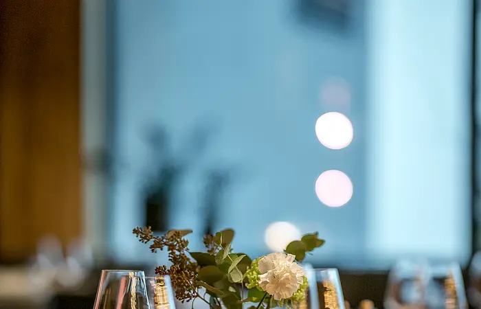 Ein Tisch gedeckt mit Wein- und Trinkgläsern, einer Vase und Servietten. Der Hintergrund ist nur schemenhaft zu erahnen.