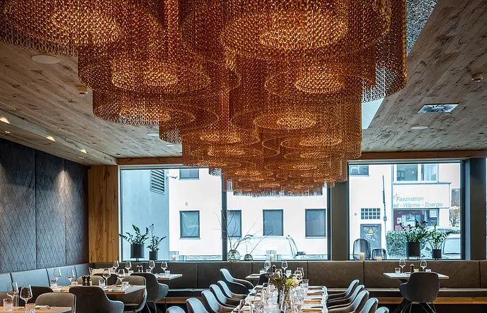 Eine lange Tafel  mit bequemen dunklen Stühlen ist aufgebaut. Im Hintergrund ist eine große Fensterfront. Von der Decke hängen braune stylische Lampen.