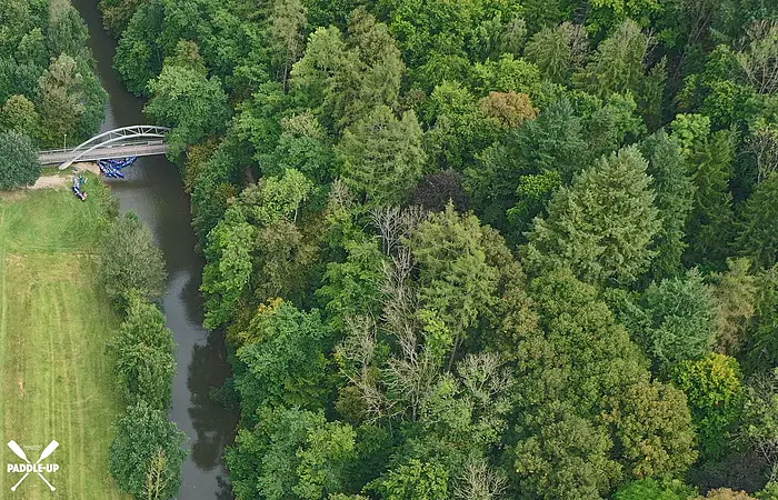 Luftaufnahme eines Flusses mit Brücke, umgeben von dichtem Wald und einer Wiese links im Bild.
