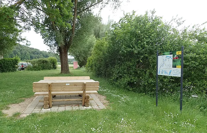 Die Sitzgelegenheit aus Holz auf gepflastertem Grund ist inmitten einer grünen Wiese am Uferrand mit Büschen und Bäumen. Daneben steht eine Infotafel zum Thema Bootwandern.