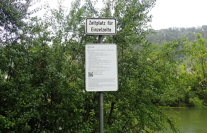 Schild mit der Aufschrift „Zeltplatz für Einzelzelte“ vor Büschen und Flussufer im Hintergrund.