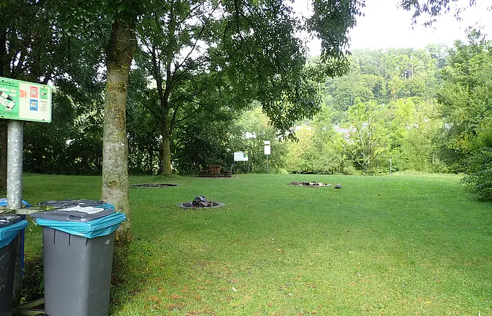 Grüne Wiese mit mehreren Feuerstellen, Bäumen und Mülltonnen links, im Hintergrund Wald und Schilder.