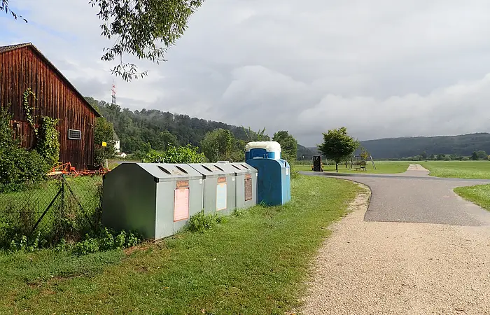 Recyclingcontainer neben einem Feldweg, links ein Holzanbau und im Hintergrund Hügel und bewölkter Himmel.
