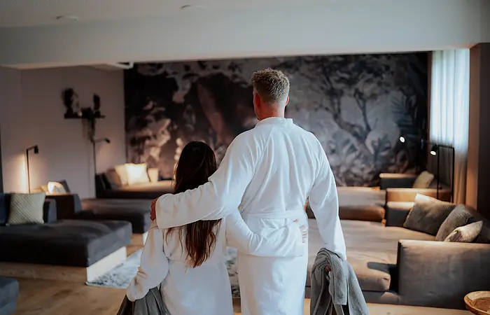 Eine Frau und ein Mann im Bademantel mit Handtücher im Ruheraum des Hotel Dirsch in Emsing. Im Hintergrund Liegen und ein Sofa mit einer kleinen Beleuchtung im Raum.