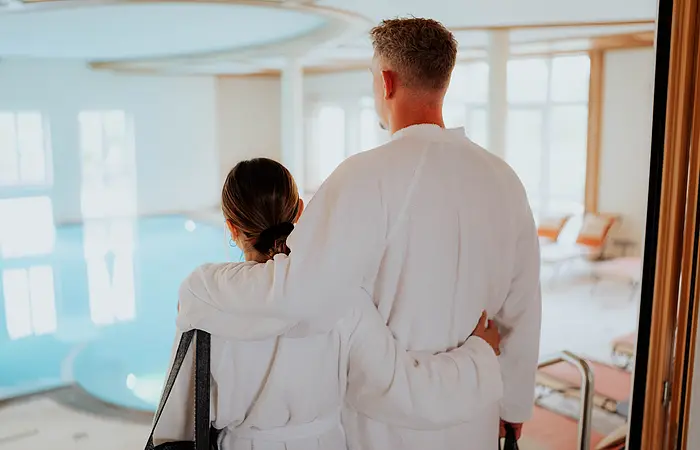 Ein Pärchen in weißen Bademäntel mit dem Rücken zu uns. Schauen auf den Indoorpool des Wellnessbereiches.