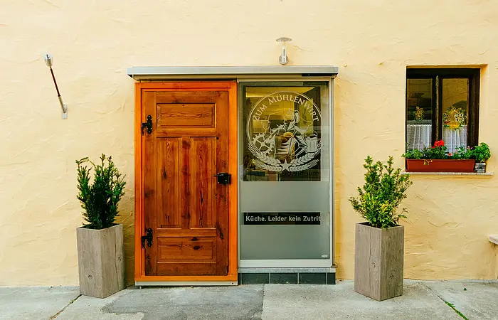 Der Eingang zum Wirtshaus ist mit einer Holztüre und daneben einer Glasscheibe mit Emblem zu finden. Dekorativ stehen davor zwei bepflanzte Blumentöpfe.
