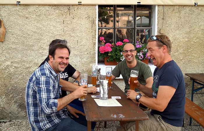 An einer Biertischgarnitur vor dem Wirtshaus sitzen vier Männer mit jeweils einem gefüllten Bierkrug in der Hand.