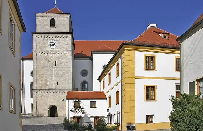 Blick auf den Eingang der Klosterkirche Heilig Kreuz. Rechts der Glockenturm in grau gehalten,   links ein Gebäude vor der Kirche mit gelben Stilelementen.