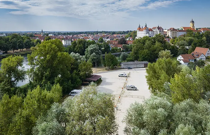Der Wohnmobilstellplatz von oben mit Blick auf das Schloss, Donau und die Altstadt von Neuburg. Der Stellplatz ist umrandet von grünen Bäumen und Büschen.