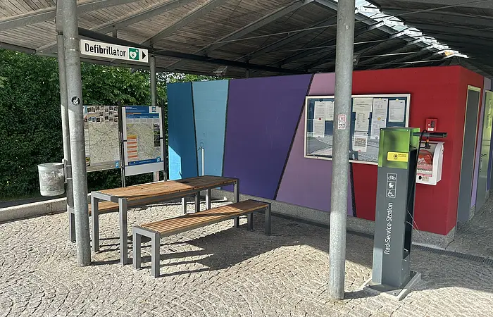 Unter dem Dach am Großparkplatz steht eine Radreparaturstation mit der man die nötigsten Reparaturen an seinem Fahrrad vornehmen kann. Links ist ein Tisch und zwei Bänke für eine kleine Rast.