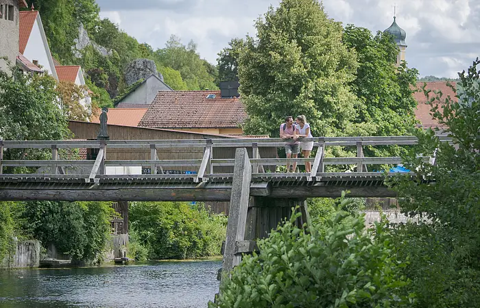 An der Balustrade der Holzbrücke vor Essing lehnt sich ein Pärchen an und schaut flussaufwärts. Hinter  ihnen sieht man einige Hausdächervon Essing umrahmt von Bäumen und deren Blätterdach.