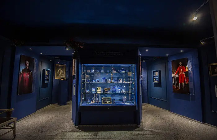 In der Mitte steht eine große Vitrine, die blau erscheint. Dahinter hängen an den Wänden rechts und links Gemälde.