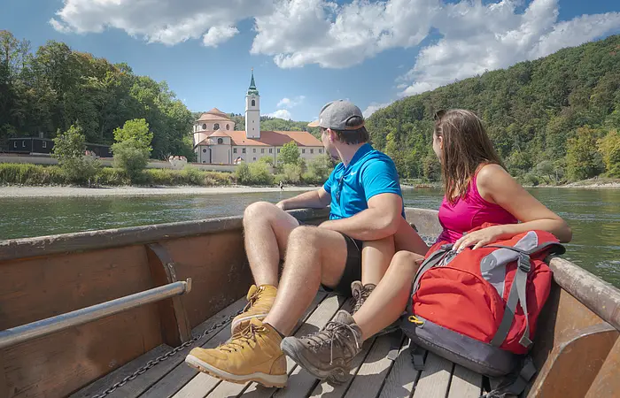 Paar sitzt in einem Boot auf einem Fluss und blickt auf ein Gebäude mit Turm und Wald im Hintergrund.