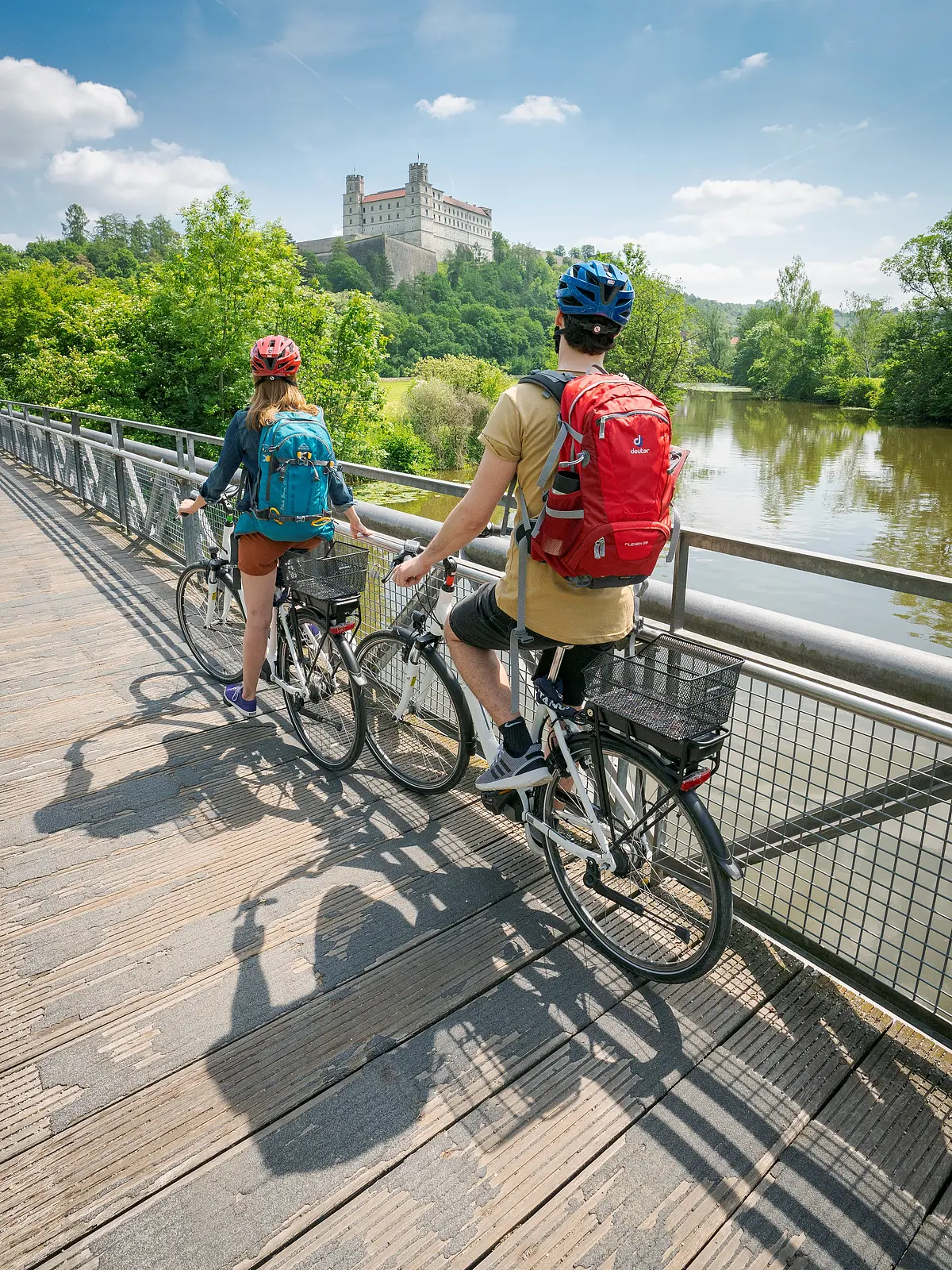 Zwei Radfahrer mit Rucksäcken und Helmen stehen auf einer Holzbrücke und blicken auf ein Schloss auf einem bewaldeten Hügel.
