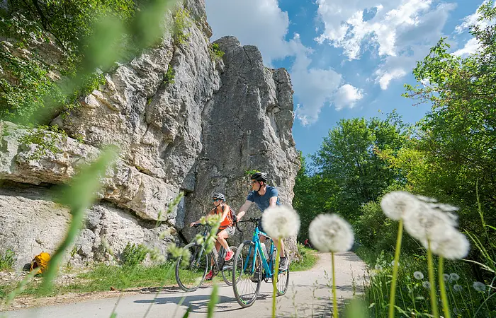 Zwei Radfahrer fahren auf einem Weg neben Felsen und grünen Bäumen bei blauem Himmel mit Wolken.