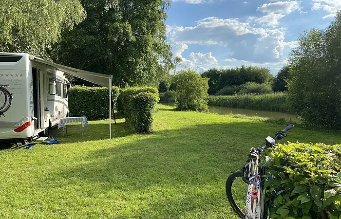 Auf dem Campinplatz steht links ein Wohnmobil mit  herausgelassener Jalousie. Hinter dem Mobil ragen große Bäume auf. Die Wiesen und Sträucher strahlen im hellen grün. Rechts lehnt ein Fahrrad an einem Strauch.