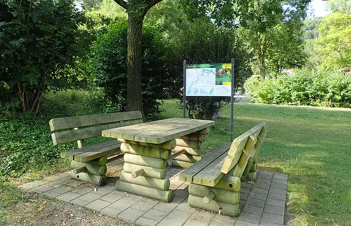 Holztisch mit zwei Bänken auf gepflastertem Platz unter Baum, im Hintergrund Infotafel und Grünfläche.