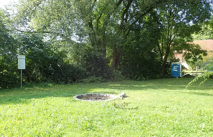 Grüne Wiese mit Feuerstelle, Bäumen, einem Hinweisschild links und einem blauen mobilen Toilettenhäuschen rechts.