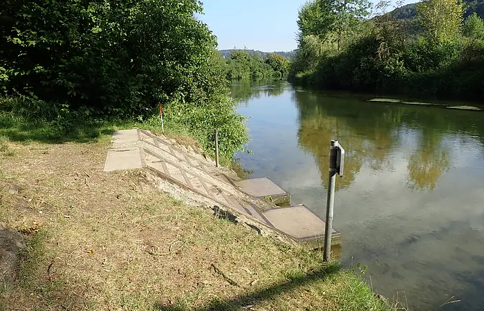 Ufer mit Stufen aus Beton an einem ruhigen Fluss, umgeben von Bäumen und Grasflächen unter blauem Himmel.