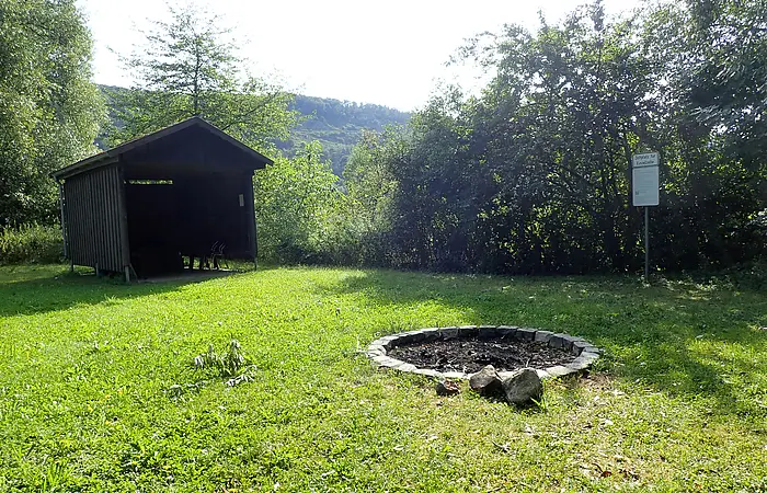Freistehende Feuerstelle aus Steinen auf einer Wiese neben einer offenen Holzhütte und einem Schild im Waldgebiet