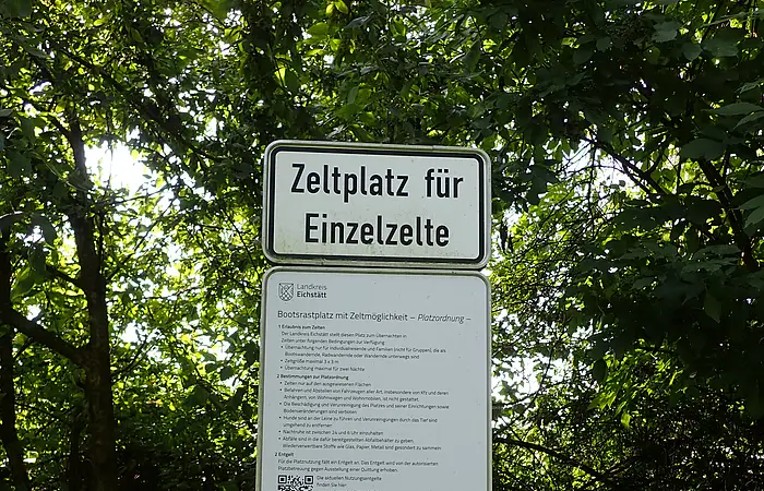 Schild mit der Aufschrift „Zeltplatz für Einzelzelte“ und weiteren Informationen vor grünem Laub.