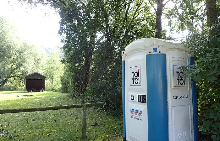 Mobiles Toilettenhäuschen mit blau-weißer Außenwand steht am Waldrand auf einer Wiese neben einem Holzzaun.