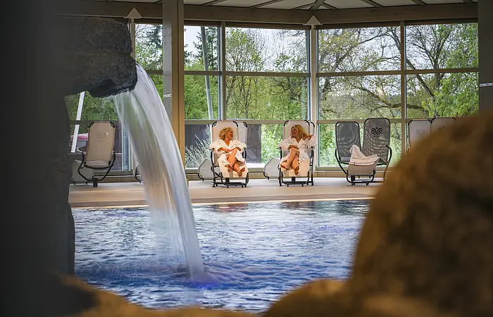 Innenpool mit Wasserfall, zwei Personen in weißen Bademänteln auf Liegestühlen, große Fenster mit Blick auf Bäume.