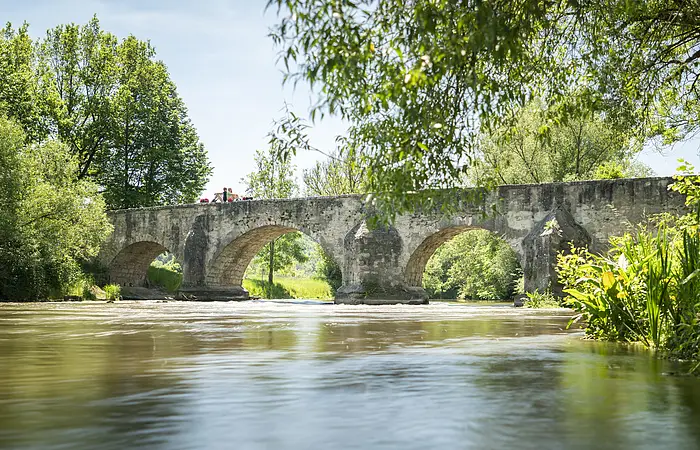 Auf der Römerbrücke sitzen zwei Wanderer Rücken an Rücken auf dem Steingeländer der Brücke. Eingerahmt von Bäumen und unter ihnen die Altmühl.