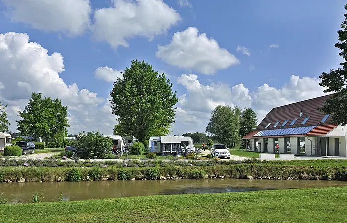 Der Campingplatz mit seinem Versorgungsgebäude, dass rechts zu sehen ist. Die Anlage ist mit Wiese und Bäumen, die Schatten spenden, angelegt.