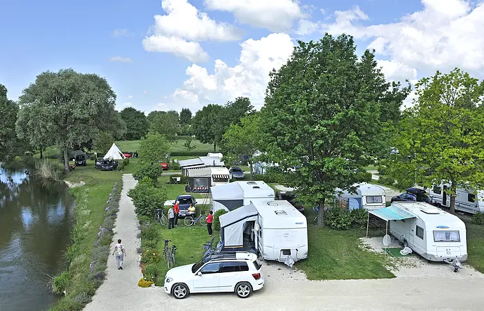 Der Campingplatz mit Wohnmobilen- und -wagen. Immer wieder gibt es Bäume, die Schatten spenden. Links ist ein Radweg direkt an der Donau.