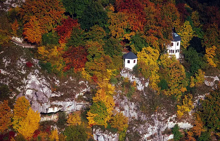 Das Gebiet der Tropfsteinhöhle von oben fotografiert. Die zwei Gebäude blitzen durch das bunte Herbstkleid des Waldes durch.