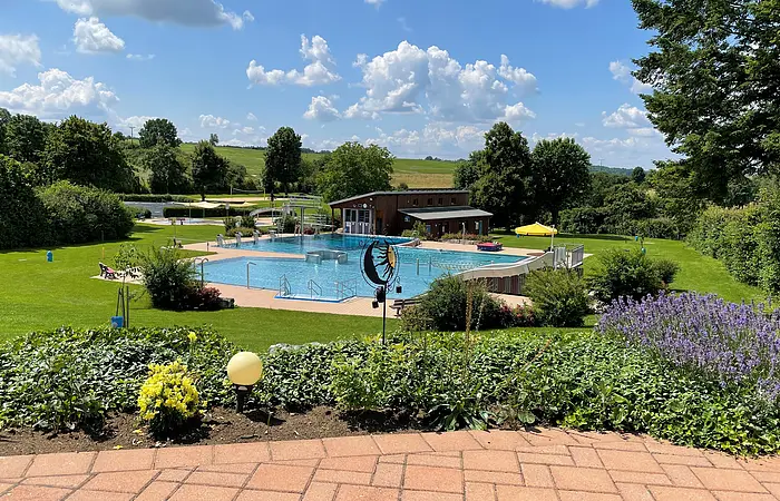 Das Schwimmbecken ist umgeben von grünen Wiesen und einigen Bäumen, die Schatten spenden. Dahinter ist ein Holzhaus zusehen. Der Bereich davor ist gepflastert.
