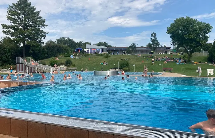 Im Freibad tummeln sich Erwachsene, Kinder im Schwimmbecken. Das Wasser schimmert hellblau.