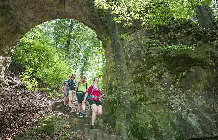 Vier Wanderer gehen einen Waldweg mit Steintreppe unter einem natürlichen Felsbogen hinab.