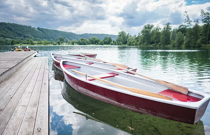Zwei Ruderboote sind an einem Holzsteg festgemacht, umgeben von einem See und bewaldeten Hügeln.