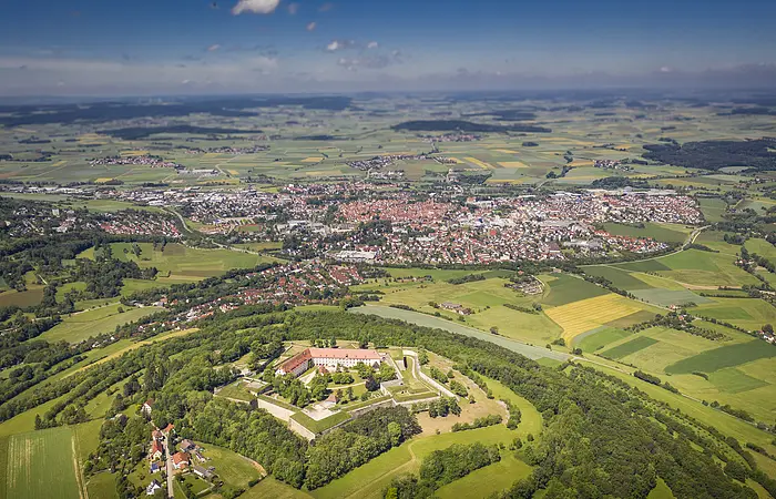 Die Wülzburg mit der gesamten Burganlage von oben. Sie ist komplett umgeben von grünen Gras und Bäumen. Dahinter kann man die Stadt Weißenburg erkennen und den himmelblauen Himmel.