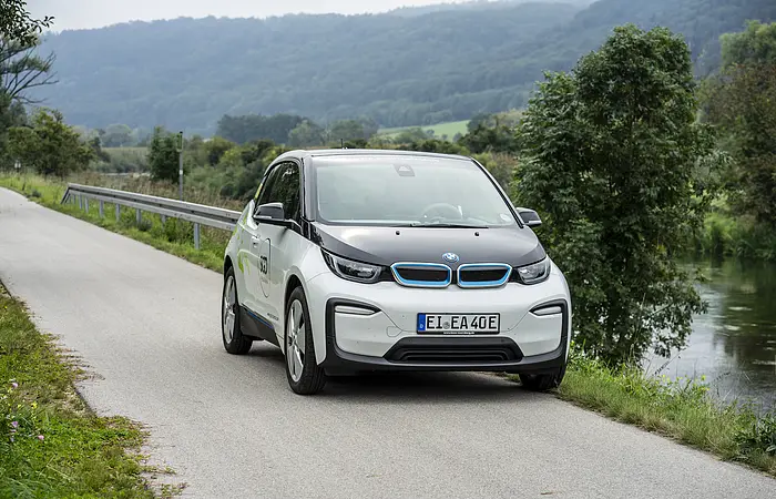 Ein weißes Elektroauto fährt auf einer ländlichen Straße entlang eines Flusses, umgeben von Bäumen und Hügeln.