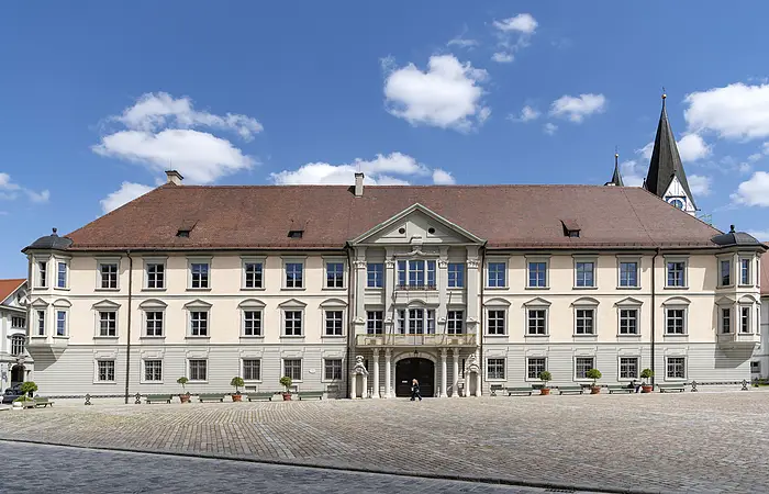 Über das ganze Bild erstreckt sich die Vorderseite der dreistöckigen Fürstbischöflichen Residenz. Der mittlere etwas vorgezogene Gebäudeteil hat ein großes zweiflügeliges Tor, das links und rechts von zwei Wachhäuschen aus Stein flankiert wird. Unmittelbar darüber befindet sich ein Balkon.
Die Fassade der oberen beiden Stockwerke ist cremefarben das Erdgeschoss ist hellgrau gestrichen. Das Dach ist mit dunkelroten Ziegeln eingedeckt.