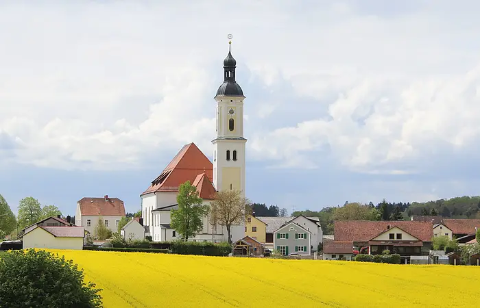 Dorf mit Kirche, Häusern und gelbem Rapsfeld unter bewölktem Himmel.