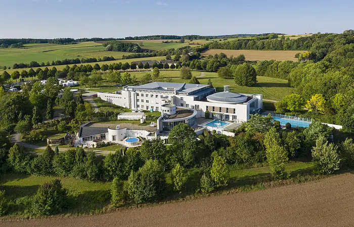 Luftaufnahme eines großen Gebäudekomplexes mit Schwimmbädern umgeben von Bäumen und Feldern.