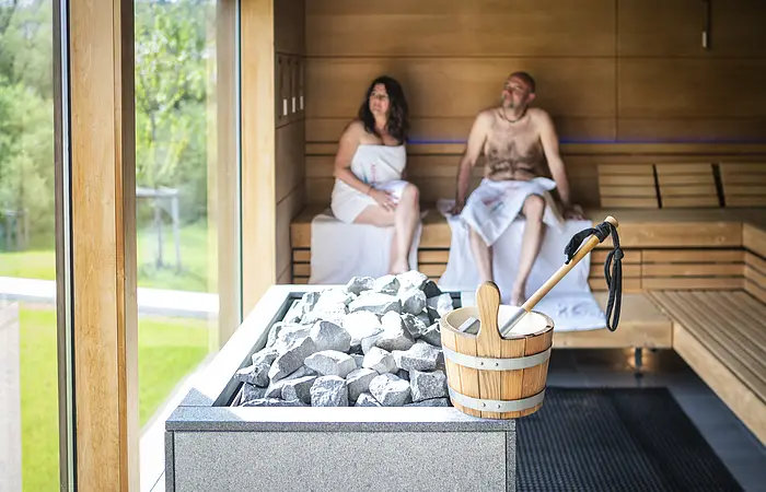 Paar sitzt in der Sauna  und im Vordergrund steht der Aufgusseimer auf dem Ofen