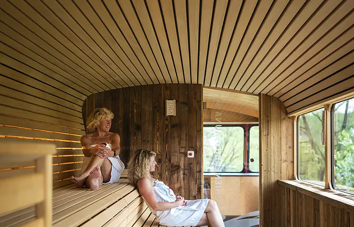 Zwei Frauen sitzen in der Schienenbus-Sauna mit dem Blick ins Freie. Im Vordergrund ist der Saunaofen